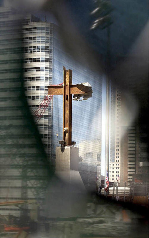 A 20-foot steel cross, mounted to a concrete foundation, inside the World Trade Center site is seen through a hole in the mesh covered fence surrounding the area. The cross was found in the rubble three days after the attack as it appears by Laborer Frank Silecchia, a construction worker clearing the debris. It was found standing almost upright and has been marked by the city as an artifact that will be part of a permanent memorial. On the right arm of the cross is attached a piece of insulation that resembles the shroud of Christ.  Photo by Mike De Sisti 8/11/02 © 2002 Appleton Post-Crescent